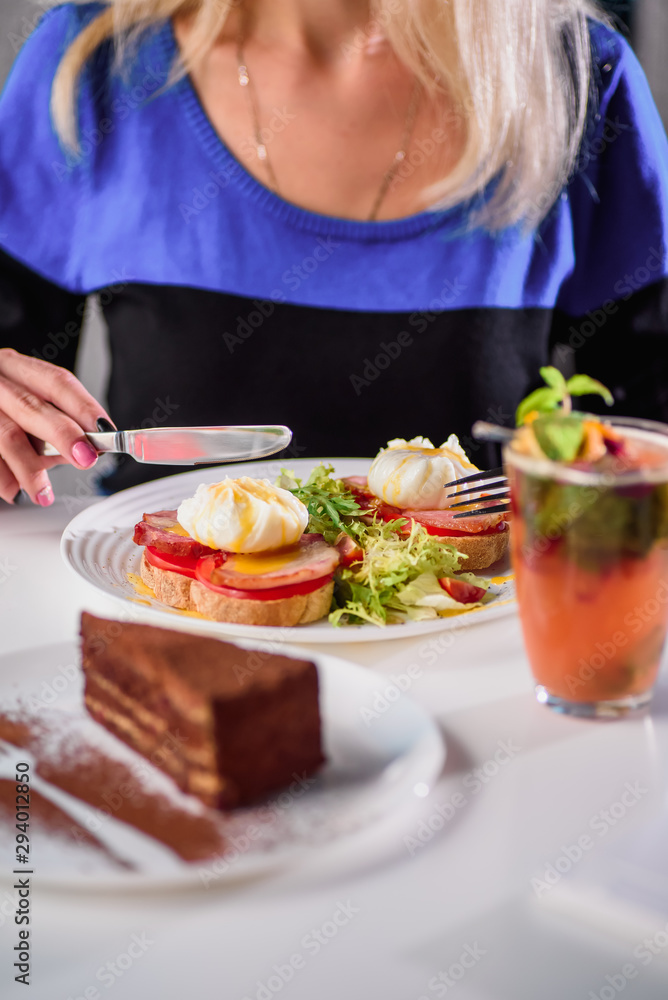 Woman eating toast with ham, tomato, poached egg and fresh salad in a restaurant. Healthy breakfast. Close-up. Space