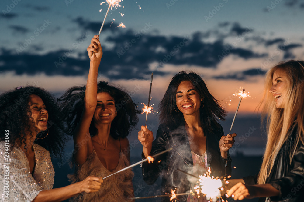 Four happy women celebrating at sunset with sparklers - obrazy, fototapety, plakaty 