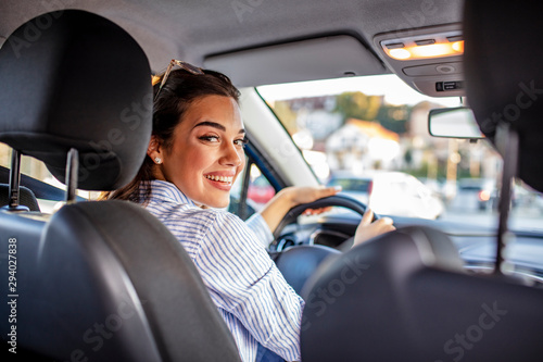 Rear view of attractive young woman in casual wear looking over her shoulder while driving a car. Woman in car indoor keeps wheel turning around smiling looking at passengers in back seat © Dragana Gordic