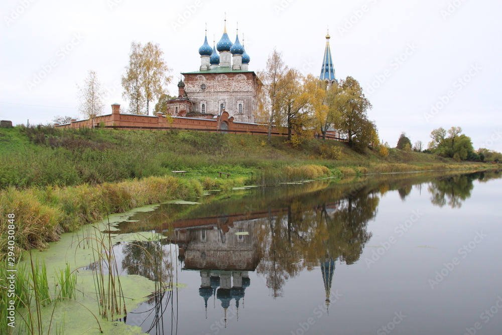 Fototapeta premium Old blue domes church and bellfray among colorful autumn trees reflecting in calm river water in cloudy day in Dunilovo village