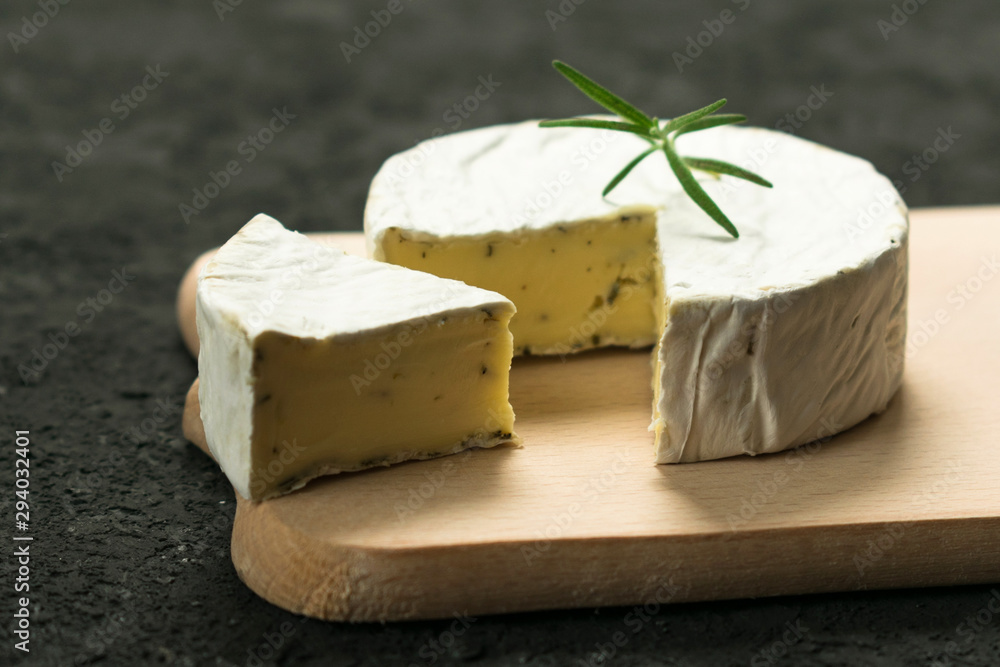 Camembert cheese with greens decorated with a sprig of rosemary on a wooden board on a black background.