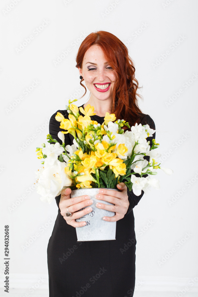 young red-haired girl in a black dress holding a vase with yellow flowers in front of her. Redhead young woman. White background. Red lips. Studio photo. Happy elegant lady