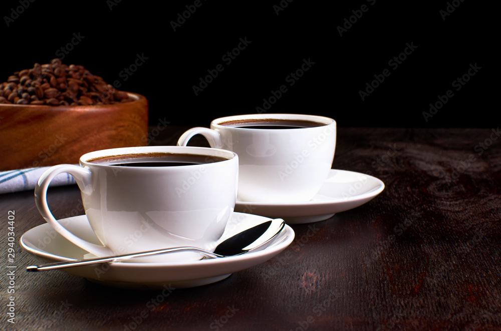 coffee on dark backgroundTwo cups of coffee and spoons in saucers on a dark wooden table. In the background, a wooden dish with coffee beans on a tea towel. coffee on dark backgroundTwo cups of coffee and spoons in saucers on a dark wooden table. In the background, a wooden dish with coffee beans on a tea towel.