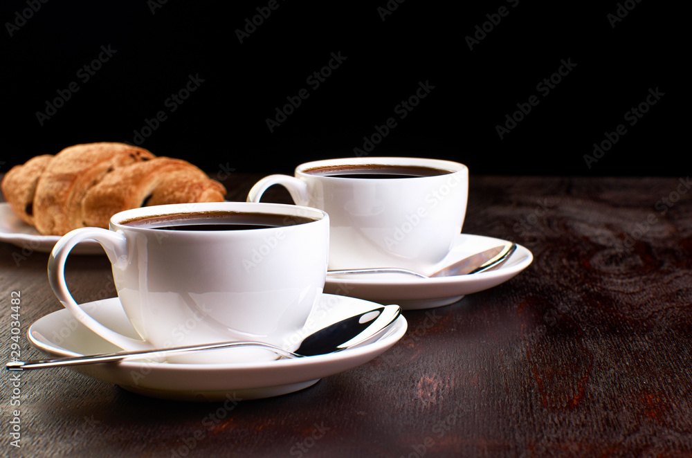 Two cups of coffee and spoons in saucers on a dark wooden table. Croissant in the background. Two cups of coffee and spoons in saucers on a dark wooden table. Croissant in the background.