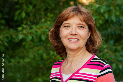 Beautiful happy European woman of middle and older age smiling, summer on background of green foliage. Portrait close-up, copy space
