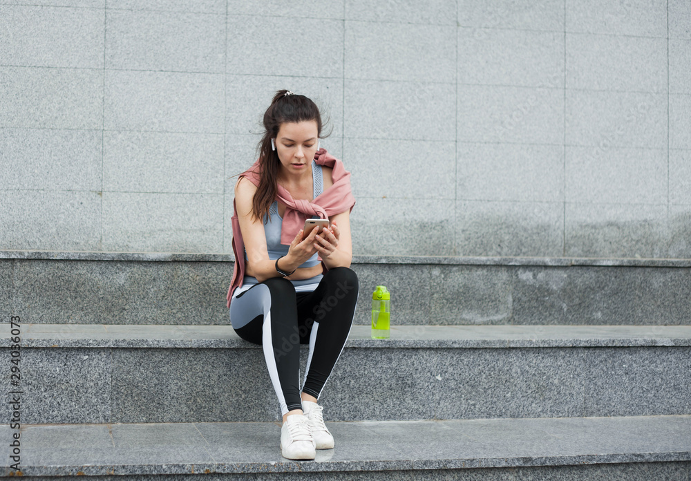 Athletic young woman in sports uniform sits, rests on the steps with smartphone and wireless headphones after a warm-up.