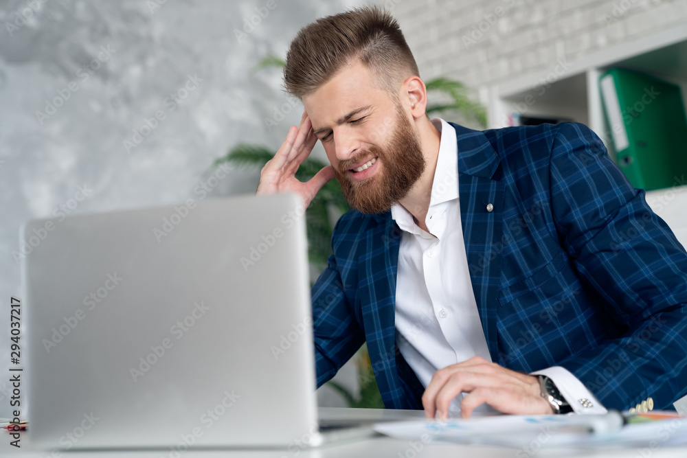 Feeling sick and tired. Frustrated young man massaging his nose and keeping eyes closed while sitting at his working place in office Feeling sick and tired. Frustrated young man massaging his nose and keeping eyes closed while sitting at his working place in office