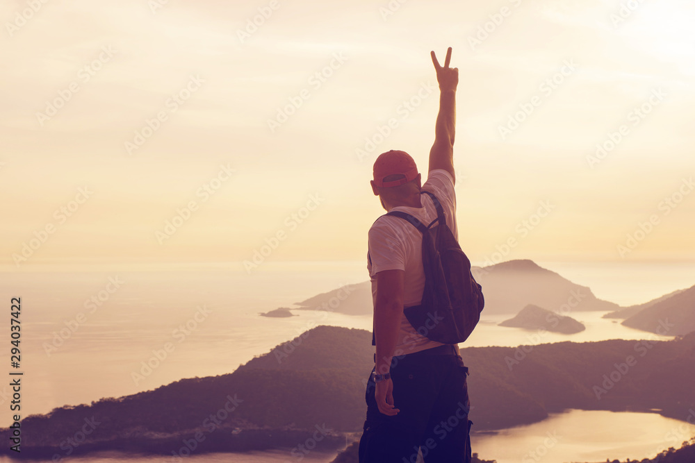 Fototapeta premium Young man standing on a hilltop with his hand raised up depicting a symbol of peace or victory, against a seascape at sunset, rear view, toned. Concept of freedom, happiness, success, victory