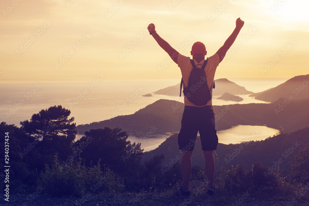 Young man with a backpack standing on top of a hill with his hands raised against the backdrop of a seascape and mountains at sunset, rear view, toned. Concept of freedom, happiness, success, victory Young man with a backpack standing on top of a hill with his hands raised against the backdrop of a seascape and mountains at sunset, rear view, toned. Concept of freedom, happiness, success, victory