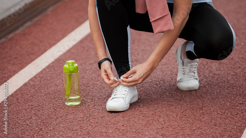 Bowed woman's hands are tying sneakers on treadmill, getting ready for a run. Next to the foot is a green sports bottle.