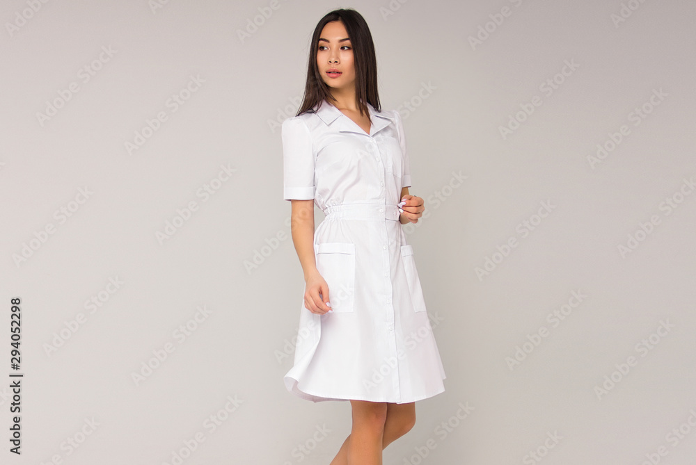 young stylish brunette nurse with long hair in white medical dress is standing and looking away on the white wall background. medical fashion concept. free space