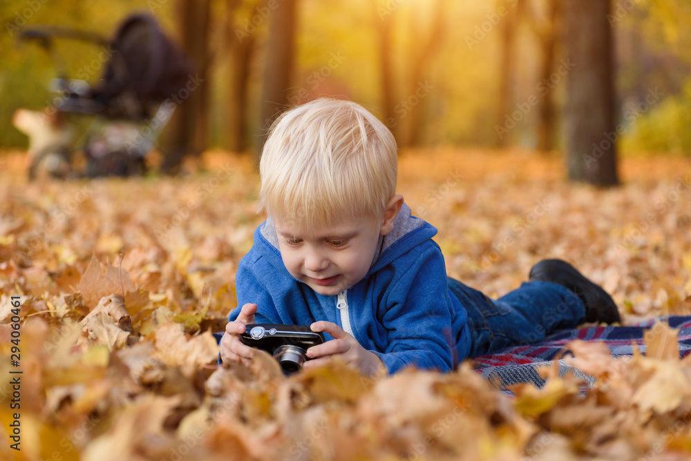 Little blond boy with camera lying on a plaid, yellow autumn leaves. Fall day Little blond boy with camera lying on a plaid, yellow autumn leaves. Fall day