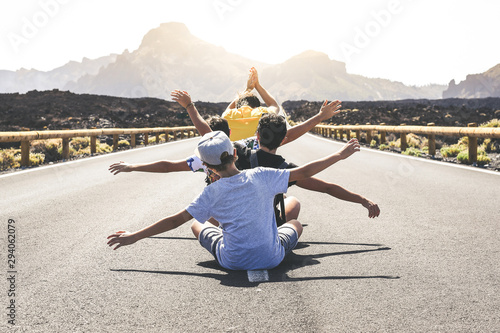 Group of teenager sitting in the middle of a long road exulting with raising arms. Back view of four teen enjoying free time on holiday with open arms. Youth, freedom, travel and adventure concept.