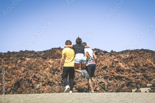 Three teenager playing happy during a school trip in Tenerife, Canary Islands. Young girl jumping on the backs of two boys walking on the sand with lava rocks in background. Youth togetherness concept