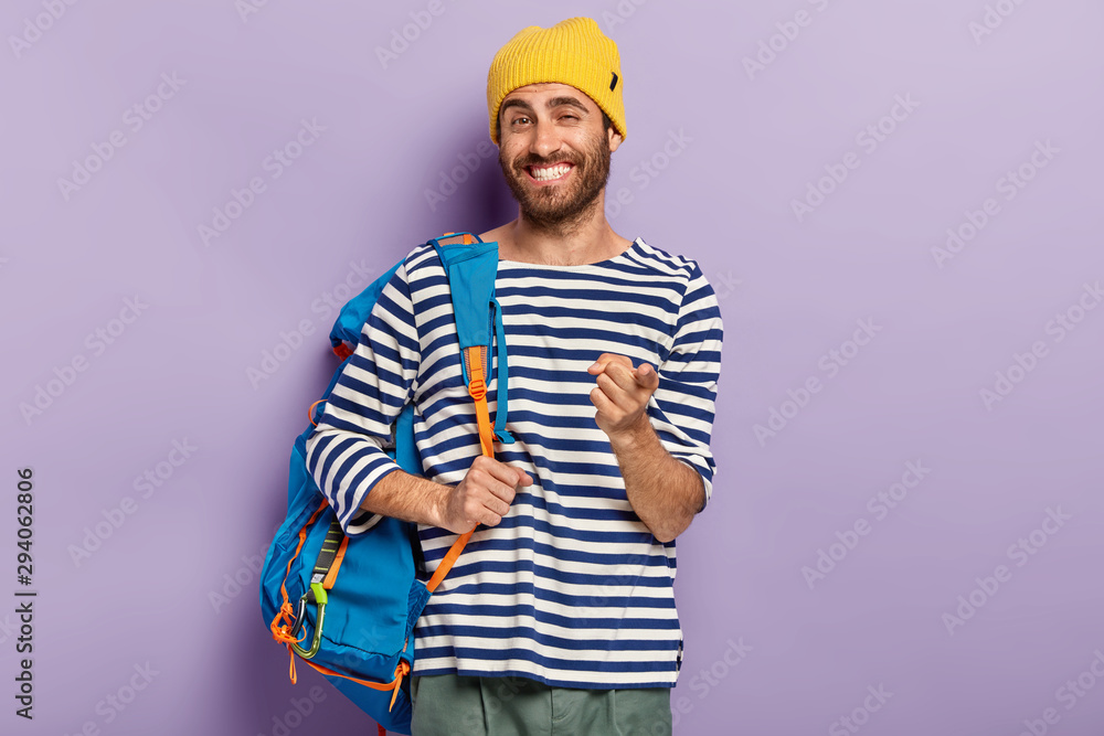 Photo of glad smiling man traveler points at you with index finger, carries rucksack with necessary things, wears yellow hat and srtiped sweater, exprsses choice, picks up you, isolated on purple wall Photo of glad smiling man traveler points at you with index finger, carries rucksack with necessary things, wears yellow hat and srtiped sweater, exprsses choice, picks up you, isolated on purple wall