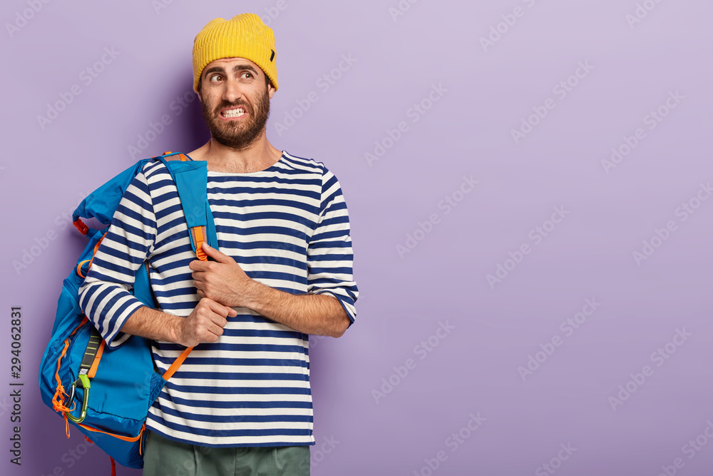 Dissatisfied young man with stubble, feels tired after long walk on foot, wears yellow headgear and sailor sweater, clenches teeth, looks unpleasantly, poses against purple wall, blank space on right Dissatisfied young man with stubble, feels tired after long walk on foot, wears yellow headgear and sailor sweater, clenches teeth, looks unpleasantly, poses against purple wall, blank space on right
