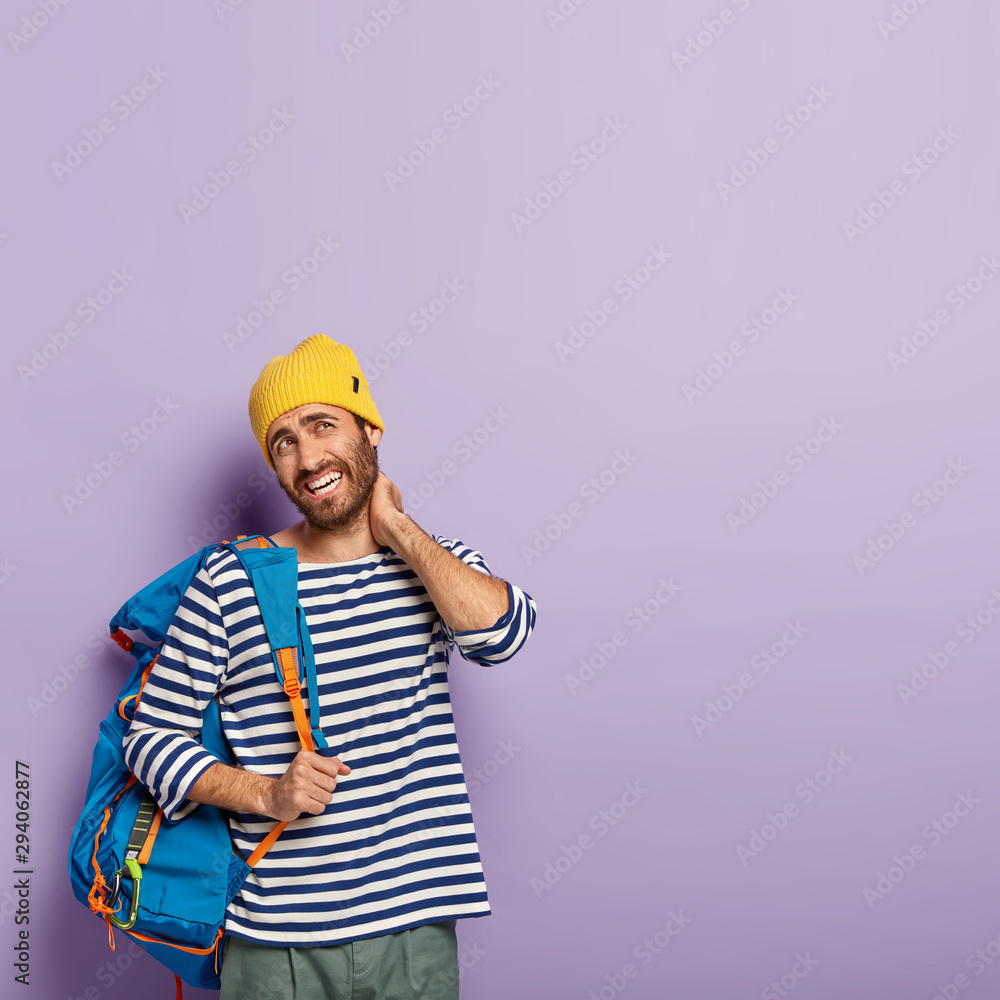 Vertical image of fatigue European man touches neck, suffers from pain in neck, dressed in casual wear, carries backpack on shoulder, smirks face, shows white teeth, isolated over purple background