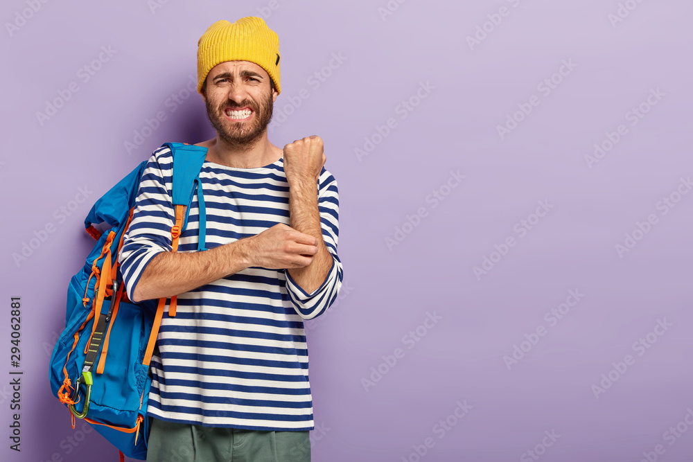 Displeased man scratches itch hand, suffers from dermatology problem, dressed in casual clothes, has travel with tourist backpack, clenches teeth, isolated over purple background, copy space area