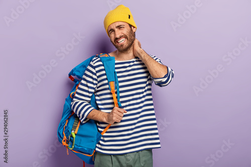 Fatigue tourist touches neck, feels stiffness, dressed in casual clothes, carries rucksack, has painful feelings, looks unhappily at camera, poses over purple background. People and tiredsome journey