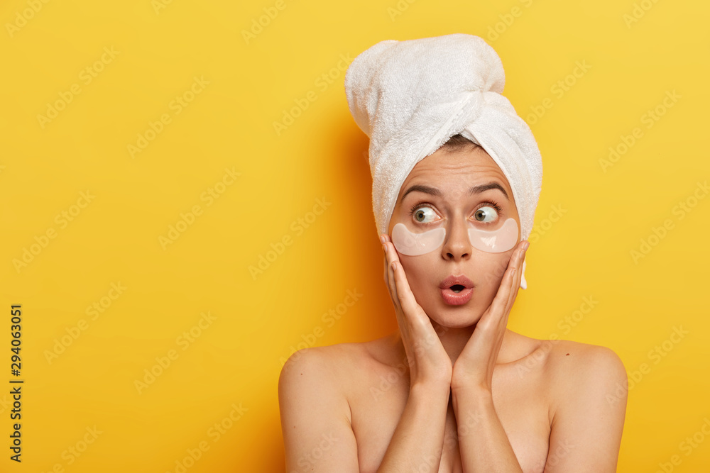 Photo of surprised young Afro woman touches cheeks with hands, looks aside with stupor, wears wrapped towel, applies white patches under eyes, models over yellow background, has spa procedure Photo of surprised young Afro woman touches cheeks with hands, looks aside with stupor, wears wrapped towel, applies white patches under eyes, models over yellow background, has spa procedure