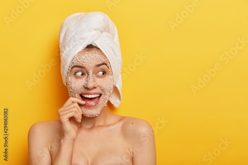 Photo of glad European girl with toothy smile, uses sea salt for spa procedures, takes shower, has smooth healthy skin, looks away, wears white towel, isolated over yellow background. Beauty concept