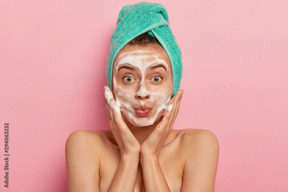 Image of attractive female washes face with foam, massages cheeks, looks surprisingly at herself, wears wrapped towel on head, removes dirt, feels freshness after taking shower, models indoor Image of attractive female washes face with foam, massages cheeks, looks surprisingly at herself, wears wrapped towel on head, removes dirt, feels freshness after taking shower, models indoor