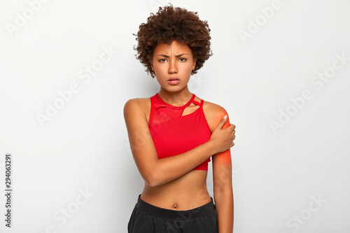 Photo of displeased Afro American woman with slim figure, dressed in red top and black trousers, touches shoulder, suffers from pain, being injured, poses against white background. Painful feeling