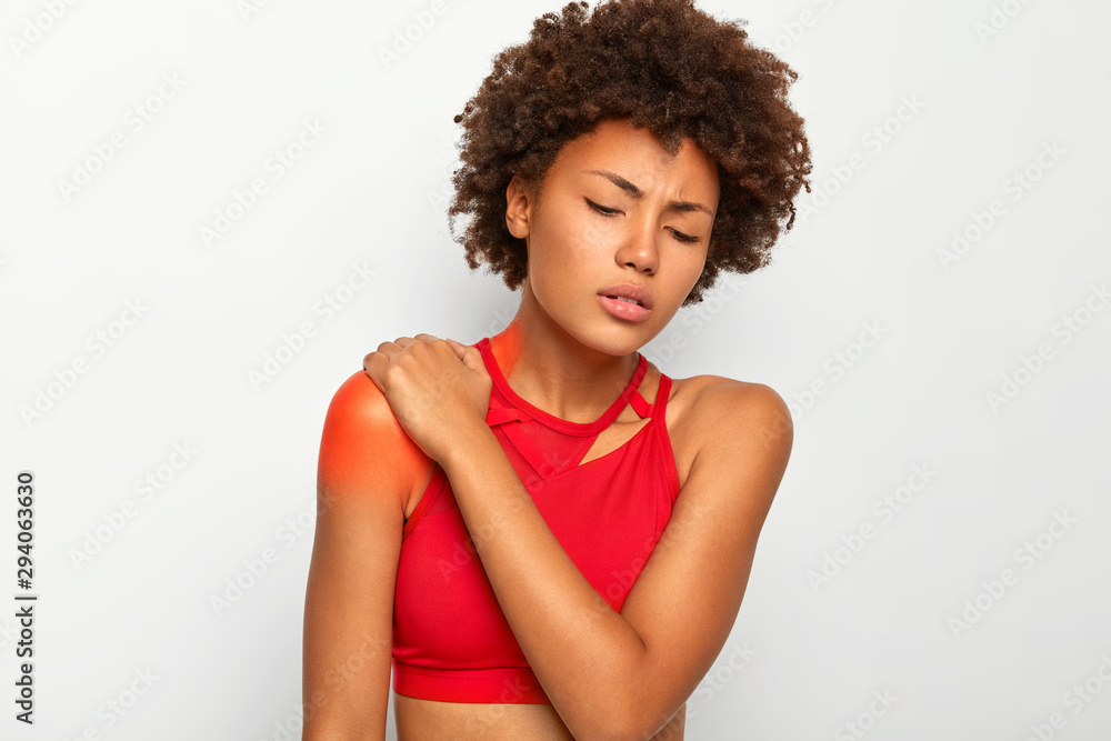 Photo of curly haired woman suffers from shoulder injury, keeps eyes closed, tilts head, touches blades, wears casual top, indicates location of pain with red spot on skin. Health problems problems Photo of curly haired woman suffers from shoulder injury, keeps eyes closed, tilts head, touches blades, wears casual top, indicates location of pain with red spot on skin. Health problems problems
