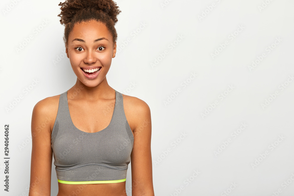 Horizontal shot of happy curly woman has bright smile, shows white teeth, dressed in casual sport bra, has fitness or workout during spare time, models indoor. Healthy lifestyle, sport and emotions