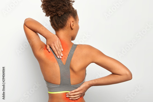Medical theme and stiffness concept. Back view of dark skinned Afro woman touches back and neck, shows inflammated zones, poses against white background, suffers from spine disease or body pain