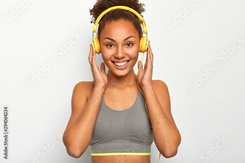 Cropped image of ethnic woman enjoys music as personal motivation, keeps both hands on headphones, smiles pleasantly, wears grey sport bra, poses against white background, has training session