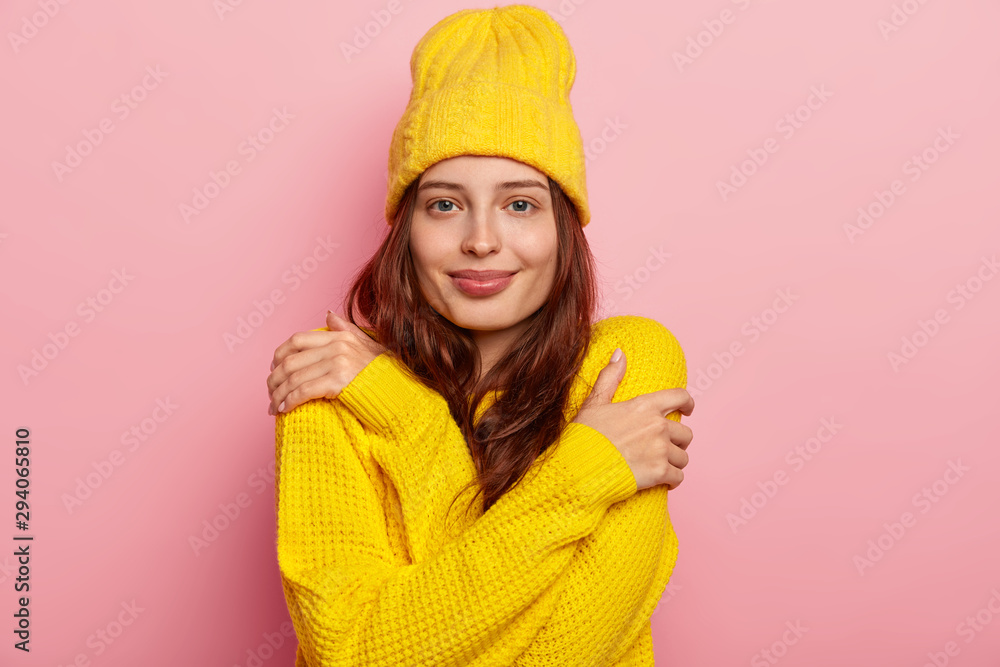 Horizontal shot of attractive young woman hugs herself, has dark long hair, tender look, wears yellow winter hat and sweater, poses against pink studio background. People and self esteem concept Horizontal shot of attractive young woman hugs herself, has dark long hair, tender look, wears yellow winter hat and sweater, poses against pink studio background. People and self esteem concept