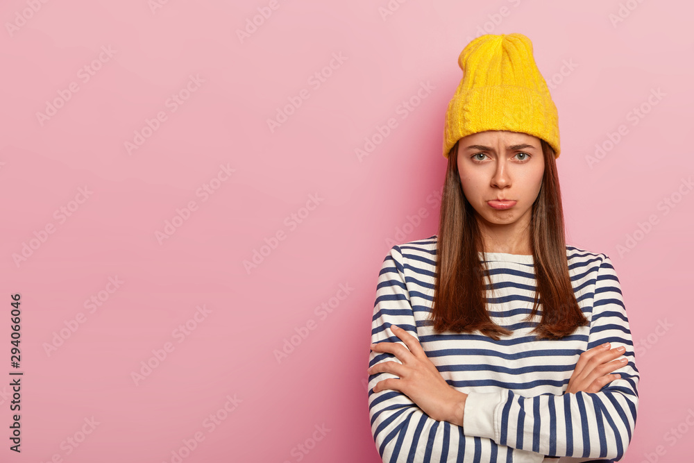 Pretty offended woman looks with sorrowful expression, frowns face and purses lips with displeasure, keeps arms folded, wears stylish yellow hat and striped sweater, models over pink wall, empty space