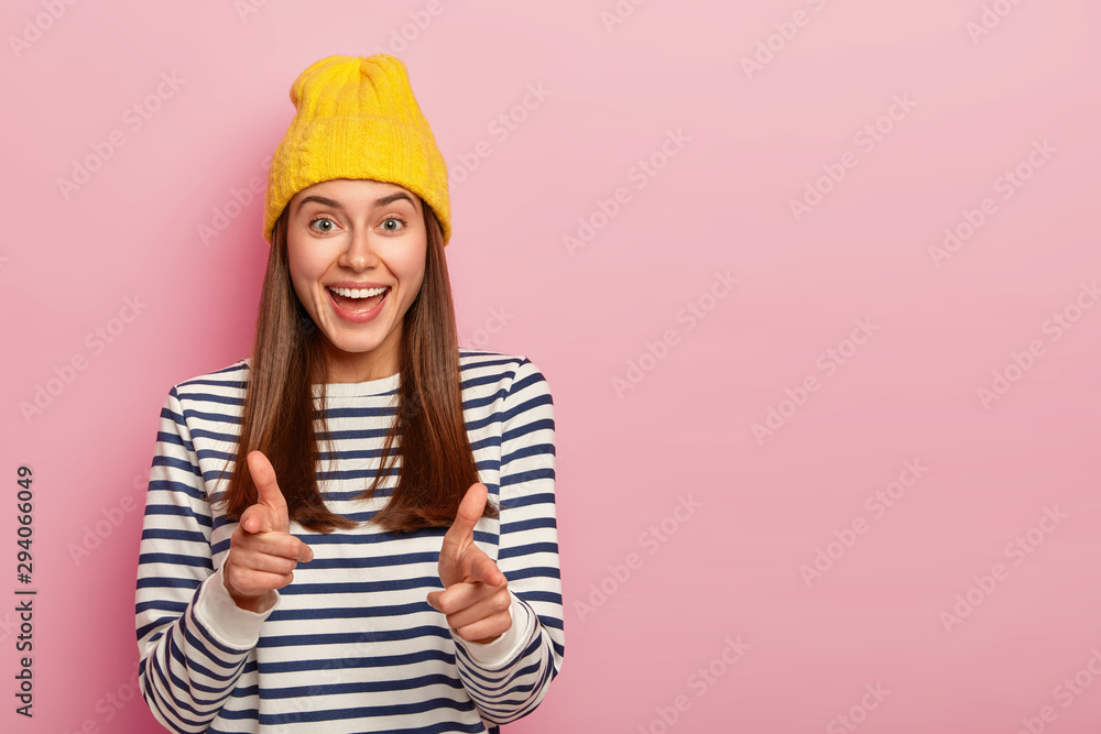 Photo of attractive young female moodel makes finger gun gesture, wears yellow hat and striped jumper, expresses choice, picks you to her team, isolated over pink background. I choose exactly you - obrazy, fototapety, plakaty 
