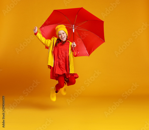 happy emotional cheerful child girl jumping and laughing with red umbrella on colored yellow background.
