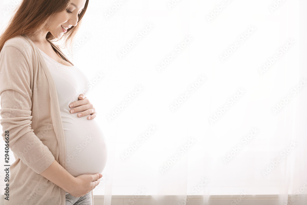 Happy pregnant woman touching her belly near window - obrazy, fototapety, plakaty 