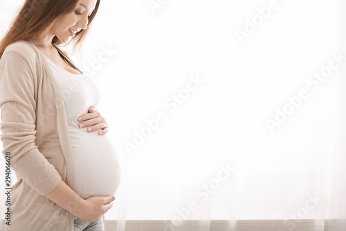 Happy pregnant woman touching her belly near window