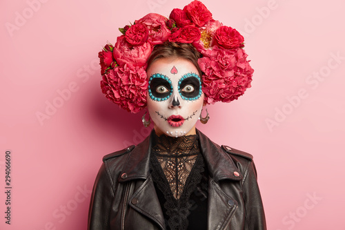 Fotografia Shocked terrified young woman has scary ghost face, wears artistic makeup for Day of Dead holiday, wears black leather jacket, models over rosy studio background
