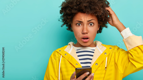 Cropped image of panicking young woman scratches head and looks with frustrated expression at camera, holds modern cellphone, thinks which answer to give on received message, wears raincoat.