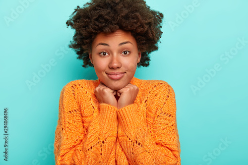Horizontal view of cheerful curious surprised African American lady with dark bushy hair  touches chin with both hands  dressed in vivid orange sweater  has natural beauty  isolated on blue wall