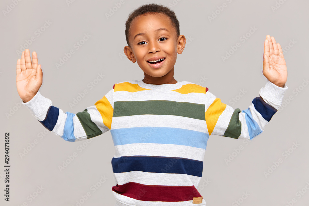 Body language. Studio shot of handsome adorable Afro American boy in striped pullover keeping hands wide apart as if holding something very large in size, measuring, having excited expression Body language. Studio shot of handsome adorable Afro American boy in striped pullover keeping hands wide apart as if holding something very large in size, measuring, having excited expression