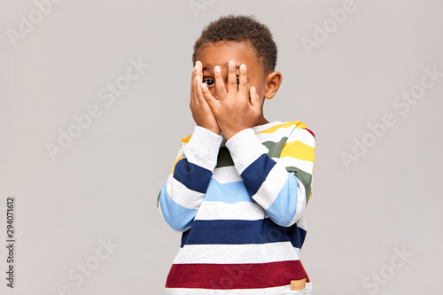 Canvas Print Frightened dark skinned little boy covering face with both hands as if being afraid to see something scary, spying through hole between fingers