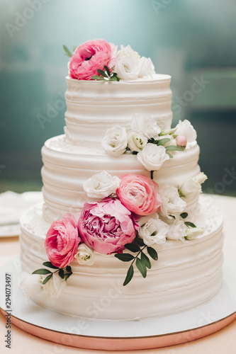 Appetizing big fresh pastry cake covered by white cream icing and decorate sweet flower serving on table. Tasty wedding event delightful dessert ready for banquet at blue light illuminate background