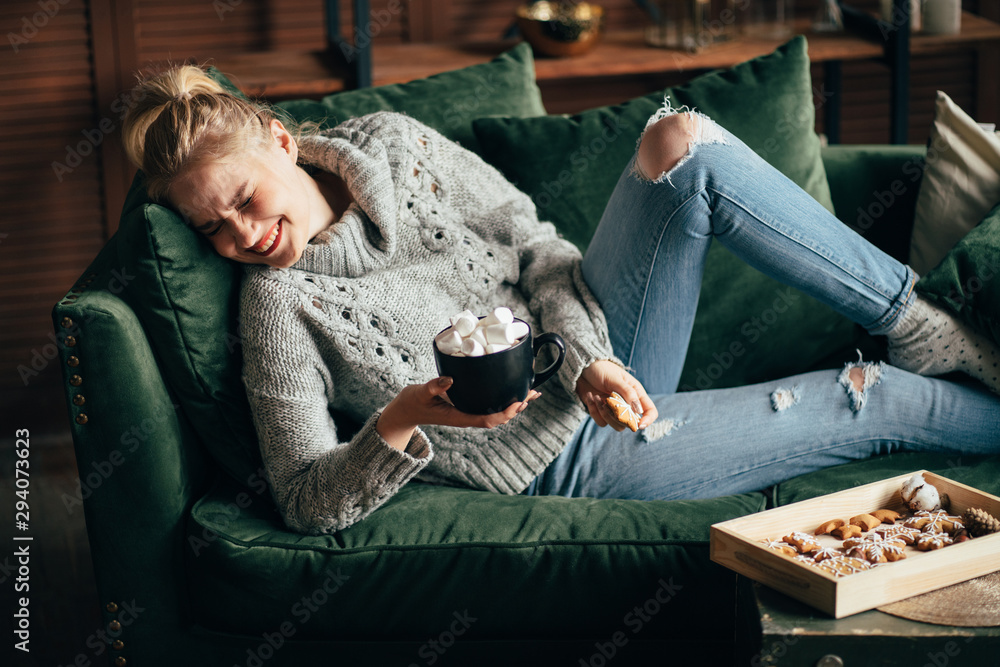 Christmas, cold autumn or winter day. Warming mood. Cheerful woman enjoy stayng at home drinking warm cocoa with marshmallows. Lazy weekend in knitted sweater on the couch. Cozy scene, hygge concept - obrazy, fototapety, plakaty 