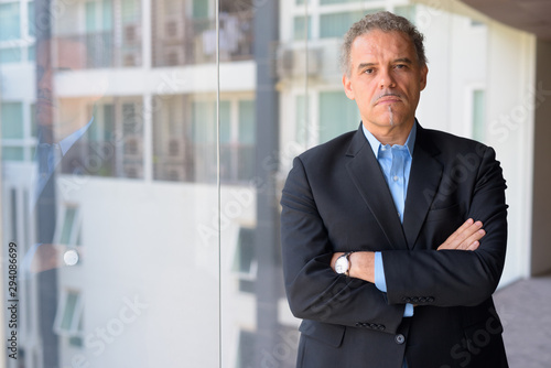 Mature Hispanic businessman with arms crossed by the glass window of office building