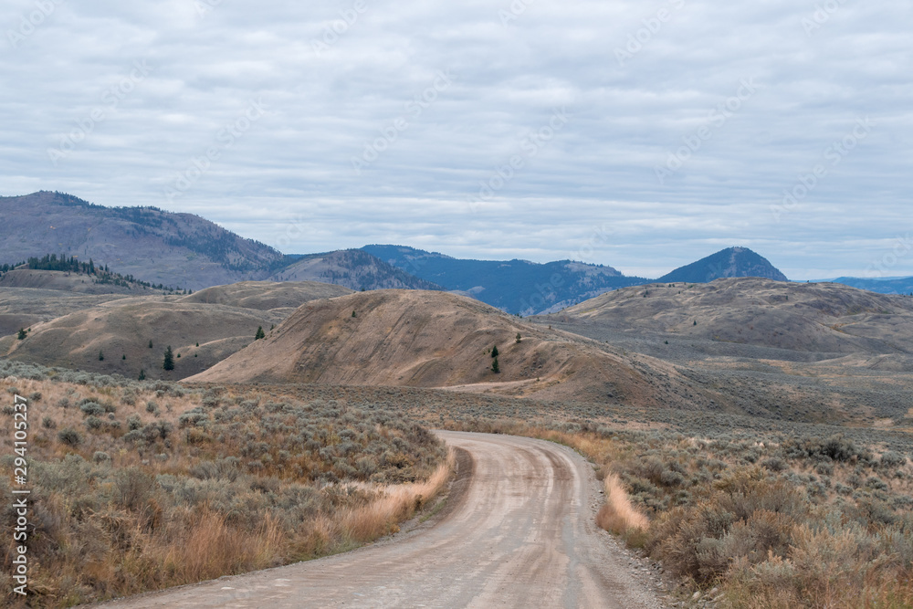 Dirt road in the valley of desert hills with shrub, dry trees, pine trees, mountain range in the background, rural countryside area, forested hills near Kamloops