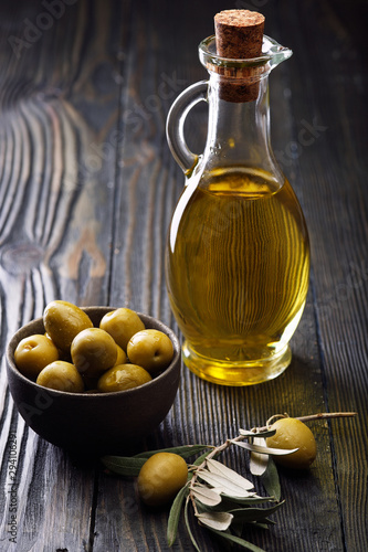 Olive oil in a glass bottle, fresh green olives in a ceramic bowl and olive branch on an old wooden table.