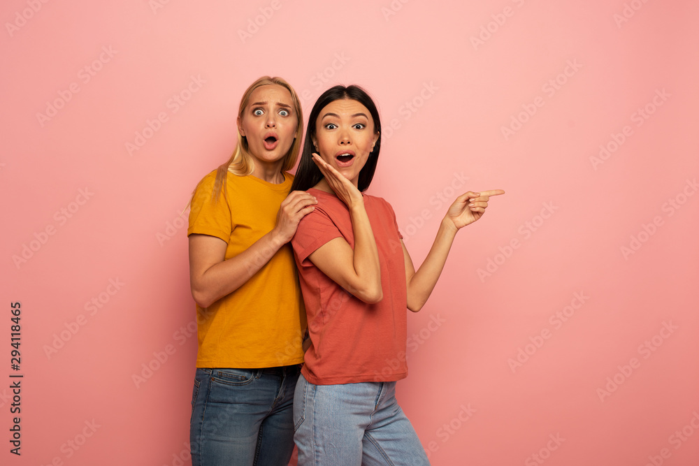 Two girls indicates something. Amazed expression face. Pink background with blank space for your text Two girls indicates something. Amazed expression face. Pink background with blank space for your text