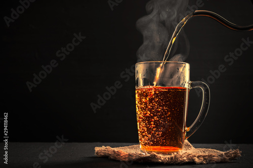 The process of brewing tea, pouring hot water from the kettle into the Cup, steam coming out of the mug, water droplets on the glass, black background