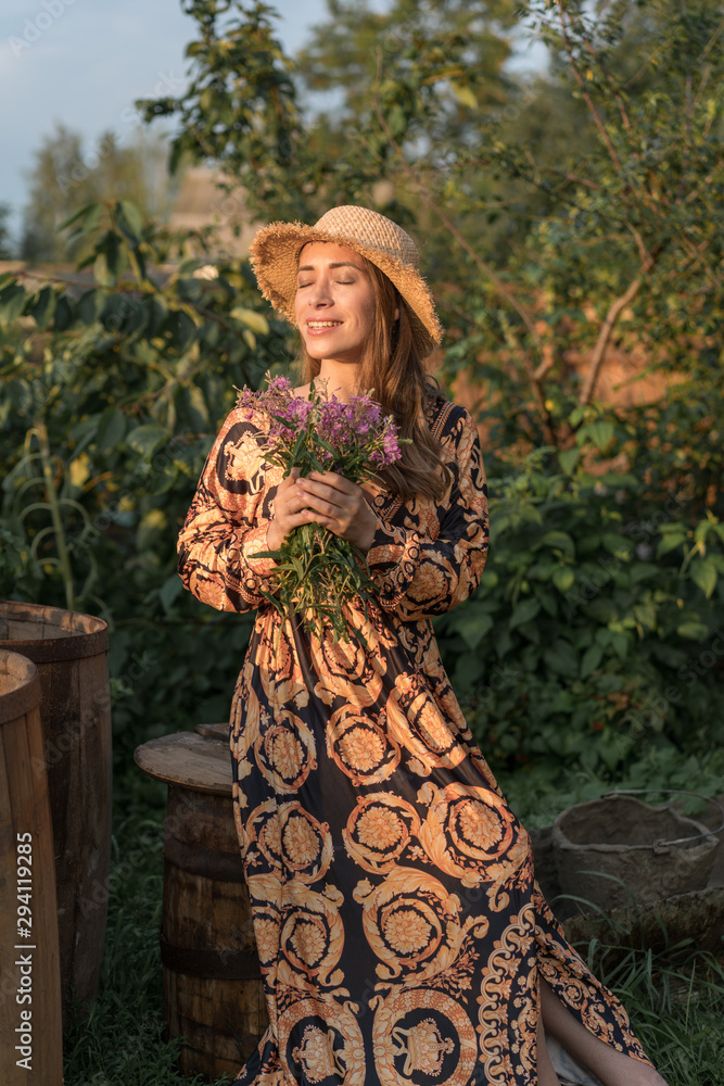 Romantic woman in golden black dress and straw village hat on sunset time holds purple flowers